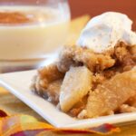 A plate of apple crisp with a dollop of whipped cream and some eggnog in the background.