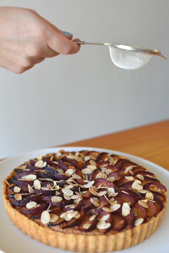 a hand holding a strainer, delicately pouring caster sugar onto a Plum and Almond Tart, adding a finishing touch to the delicious dessert