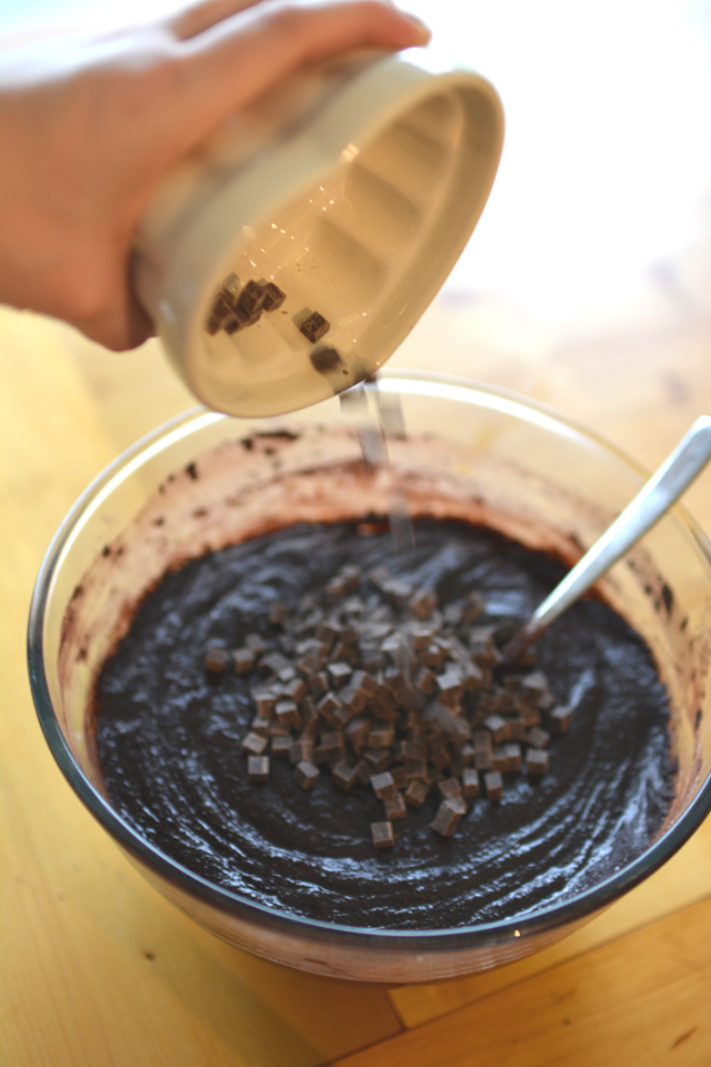 a hand pouring chocolate chips into the mixture of butter, cocoa powder, and flour