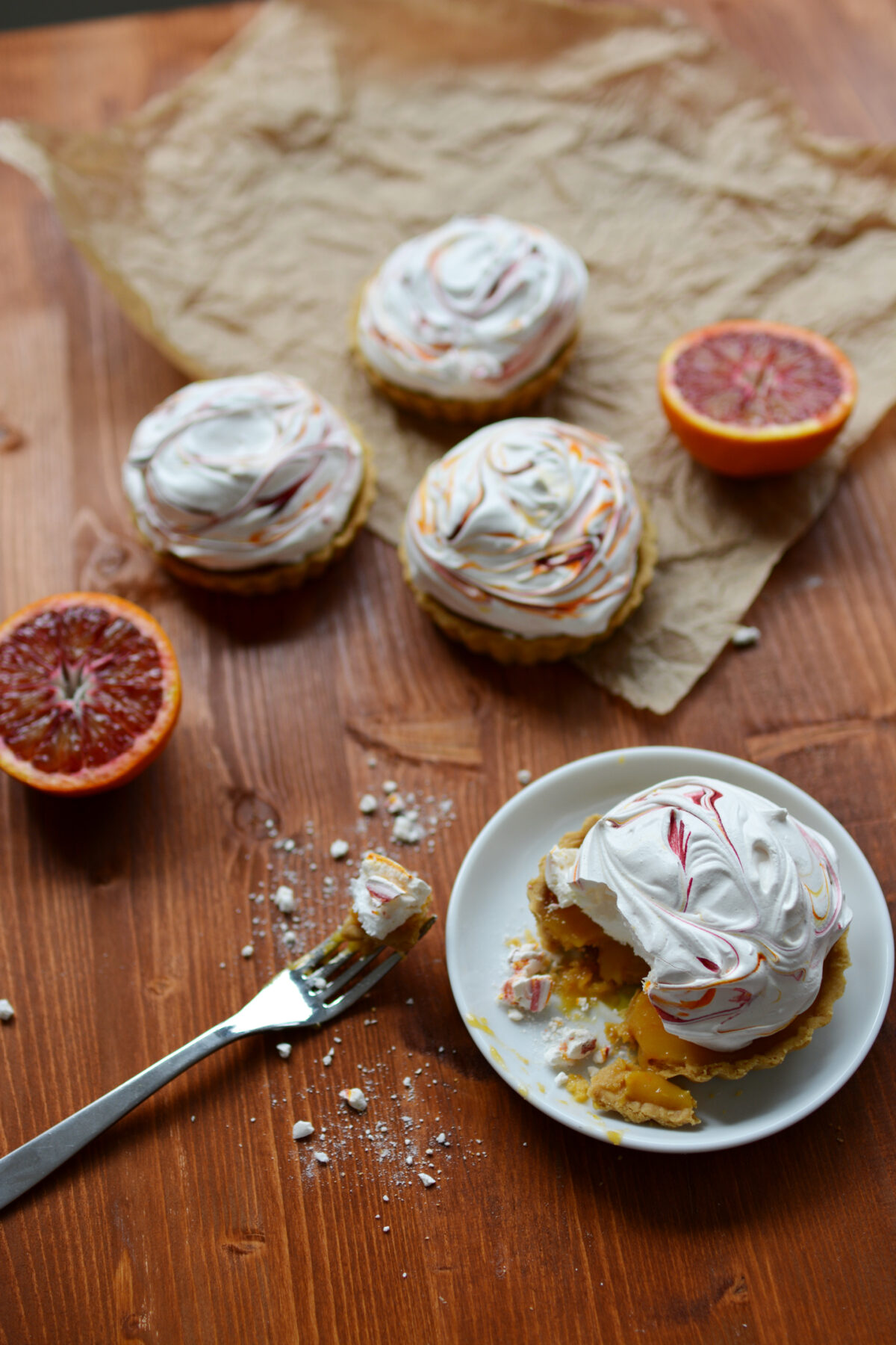 top-down view of a delightful display featuring four blood orange meringue pies on a table. one pie is positioned beneath a plate, its cut portion revealed by a fork. the trio of remaining pies is artfully presented together, enhanced by two elegant slices of blood orange