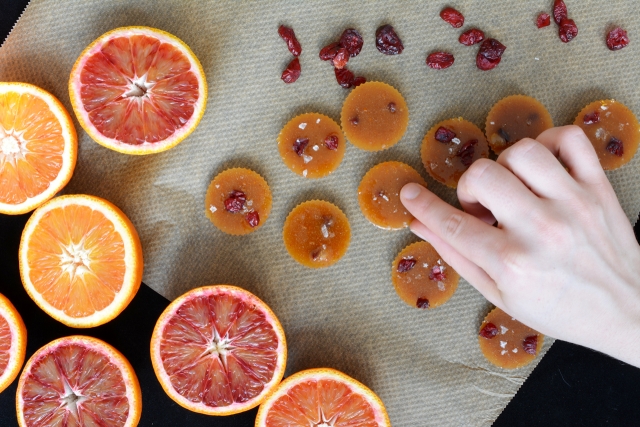 top-down view image of a hand pressing a single salted blood orange toffee from a batch, with dried cranberries nearby, and slices of blood oranges and regular oranges on the other side
