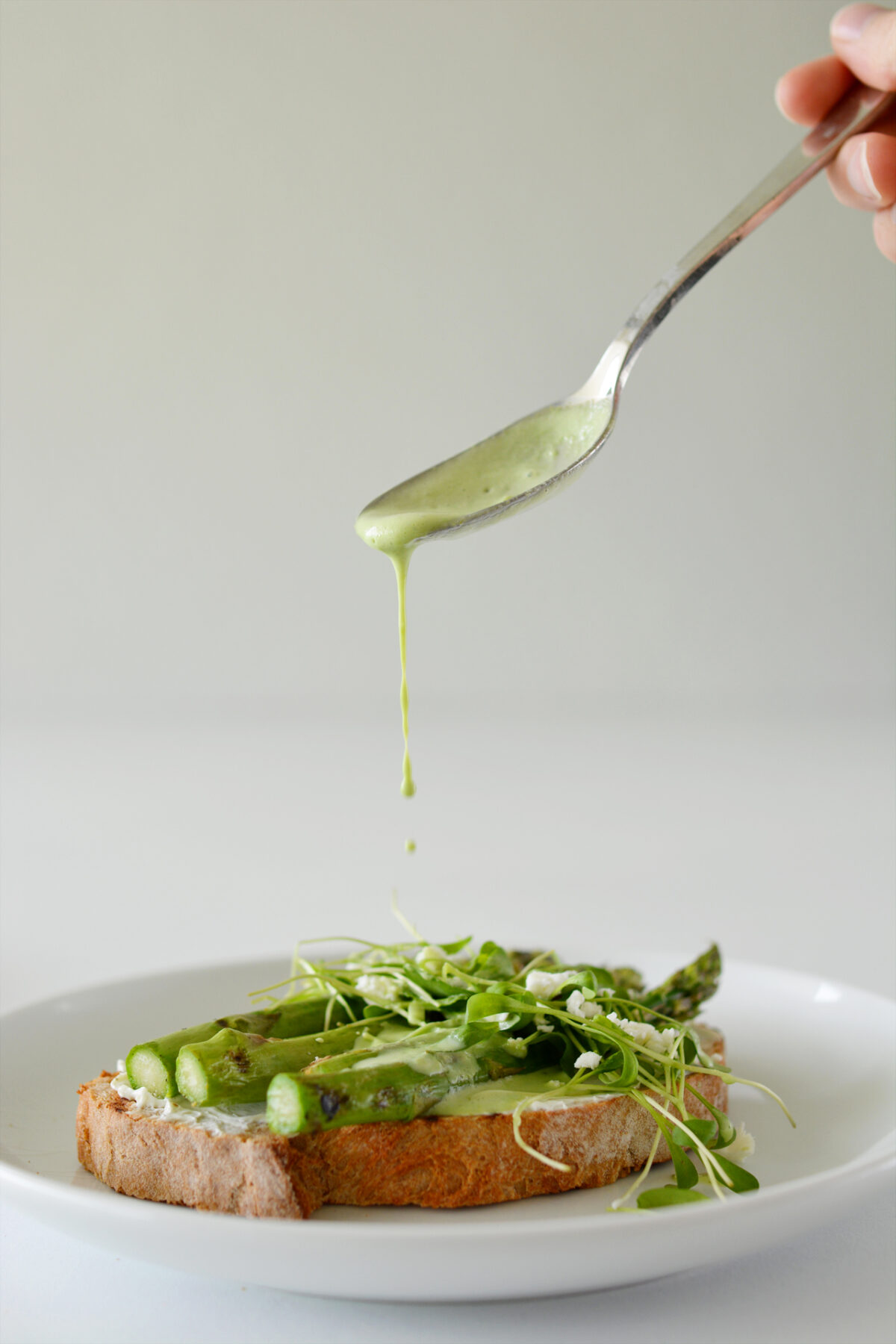 a spoon drizzling yogurt dressing over toasted bread with asparagus, parsley, cress, and feta cheese
