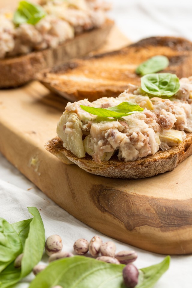 close up image of a crusty bread topped with a flavorful mix of two beans and artichoke on a cutting board with fresh basil leaves
