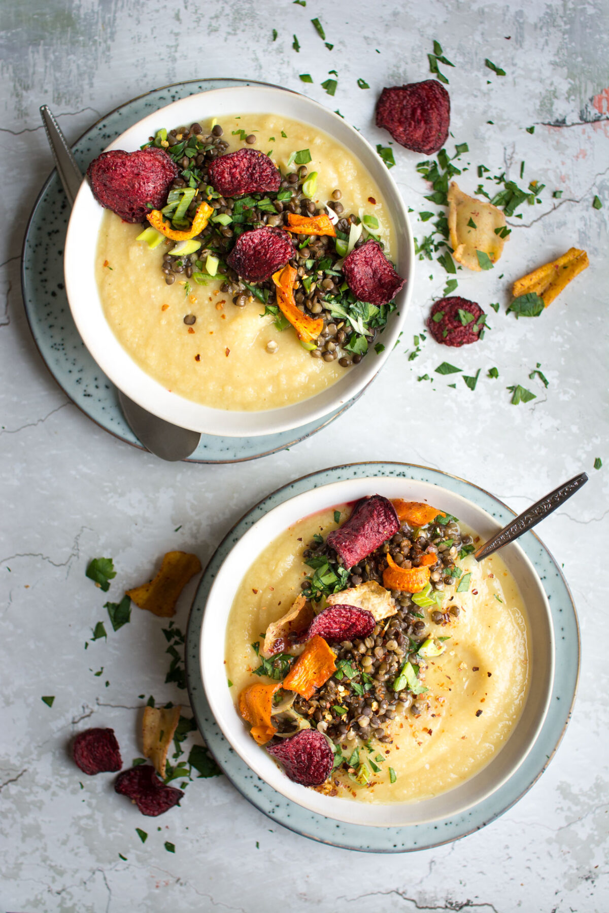 top-down image of 2 bowls full of delicious, sweet parsnip and apple soup, loaded with lentils and roasted vegetable crisps topped with black lentils, spring onion, parsley, chili flakes, and root vegetable chips 
