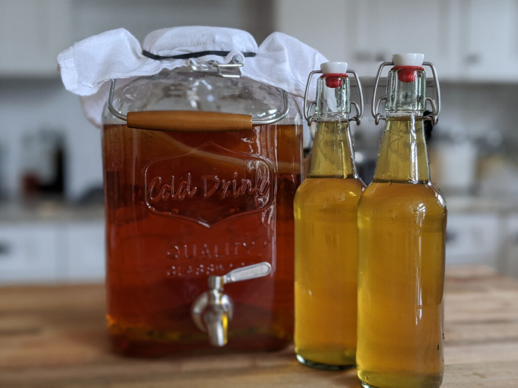 A large fermentation jar filled with kombucha along with two finished bottles of kombucha.