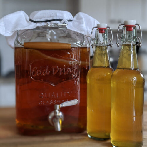 A large fermentation jar filled with kombucha along with two finished bottles of kombucha.