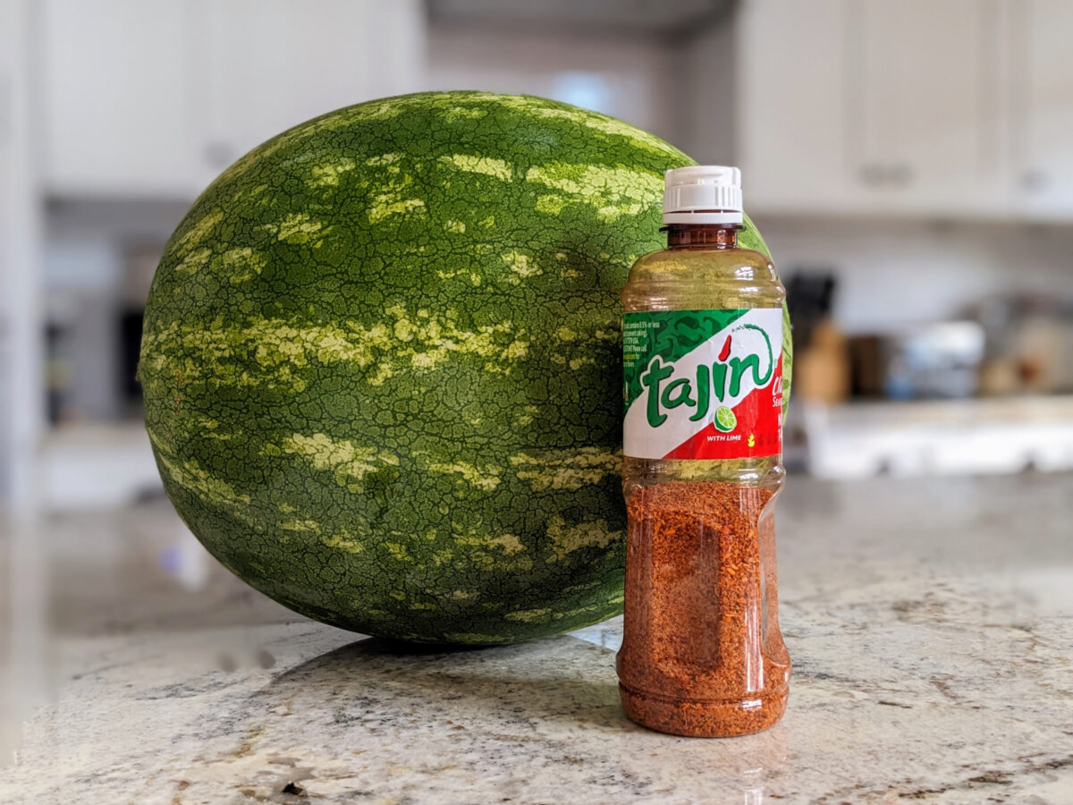 A bottle of Tajin chili lime seasoning with a watermelon behind it.