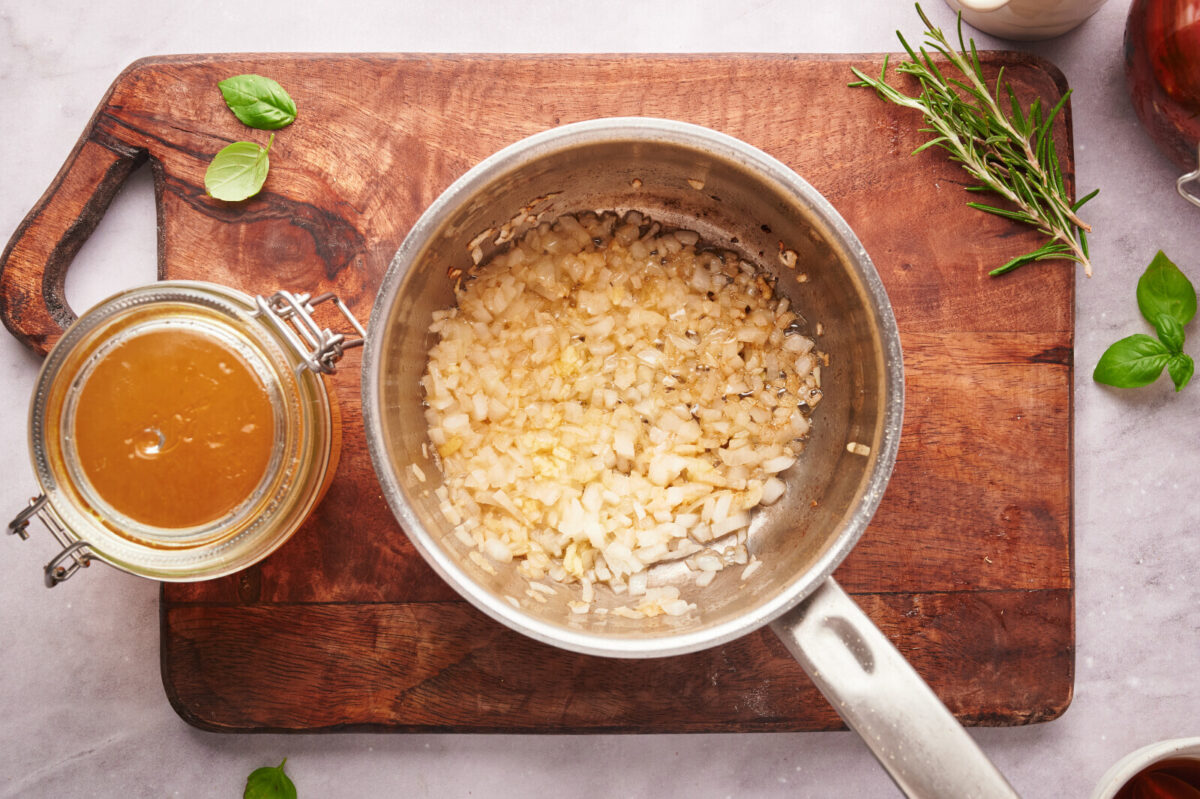 Onions and garlic simmering in butter in a pot on a wooden cutting board with a jar of vegetable stock.