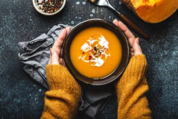 Female hands in yellow knitted sweater holding a bowl with pumpkin cream soup on dark stone background with spoon decorated with cut fresh pumpkin, top view.