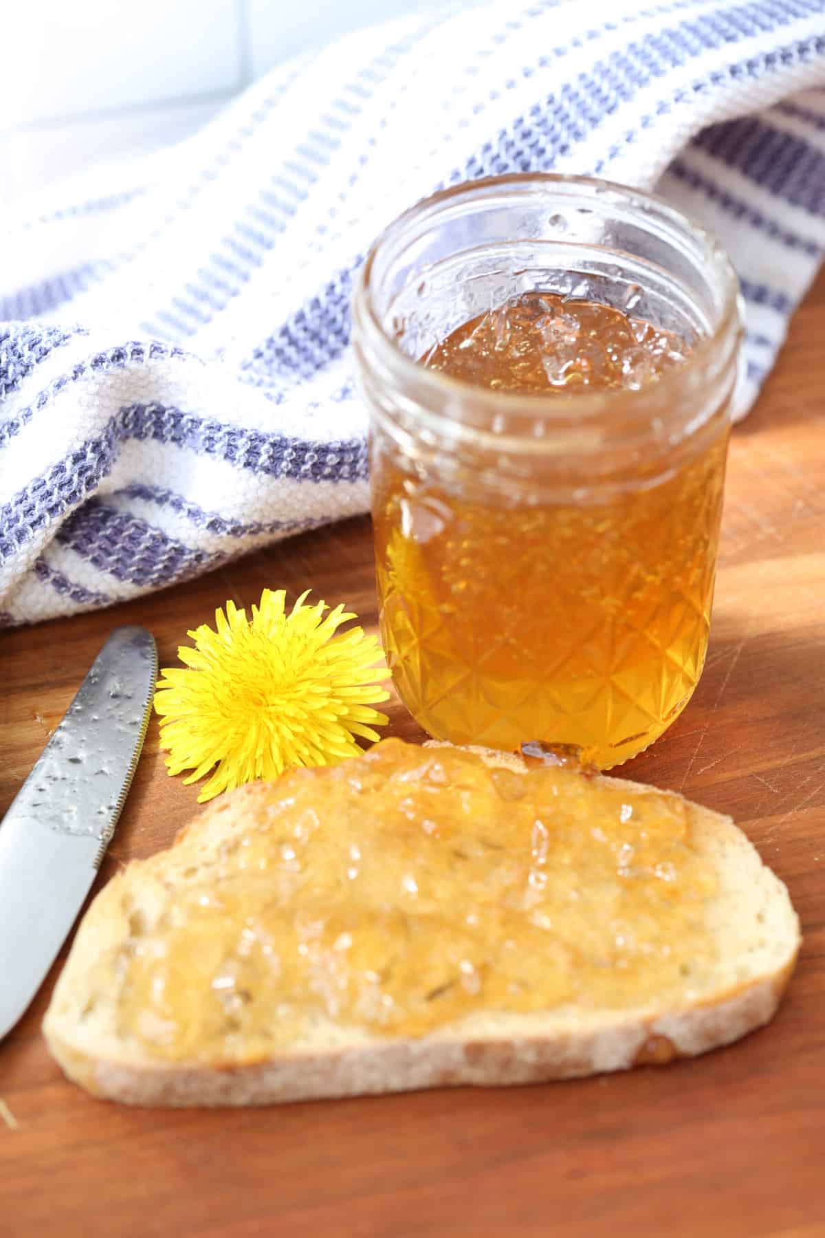 Vertical view of a jar of dandelion jelly with a dandelion flower on the side and the jelly spread on a piece of toast sitting on a wooden cutting board.
